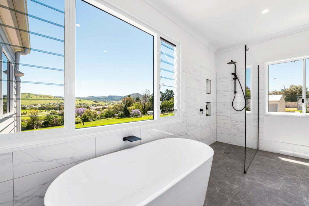 A modern master bathroom with floor to wall tiles, a standalone bath, a walk-in shower with recessed wall storage and views over the valleys of Killarney