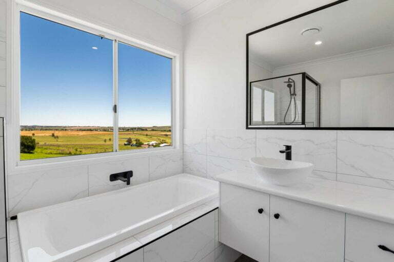 modern family bathroom with a single vanity unit, bath tub, large window overlooking acreage. Tiles are light and marbled, fixtures are black.